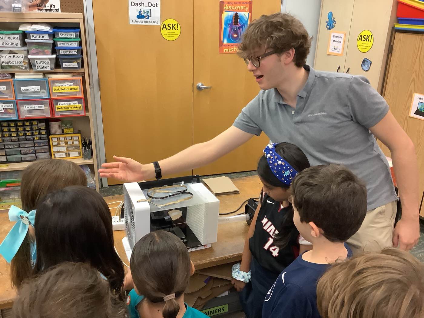 The instructor explains airflow to a group of students gathered around a fan and a rocket fin mounted on a workbench.