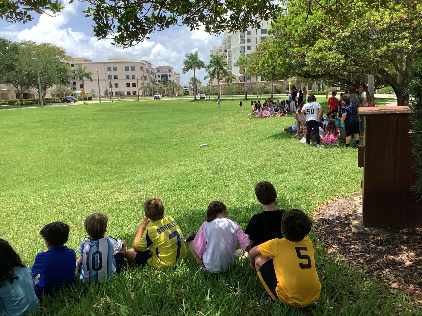 A class seated on the grass at the edge of a field, watching a model rocket climb overhead.