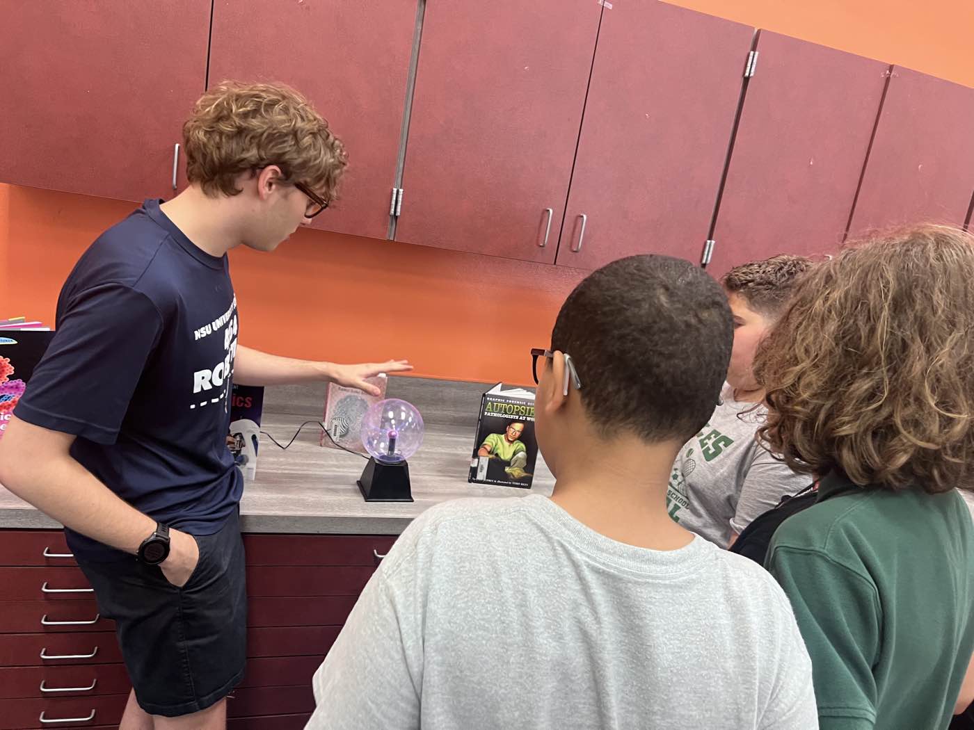 The instructor demonstrating a plasma globe to a small group of students gathered around a counter.