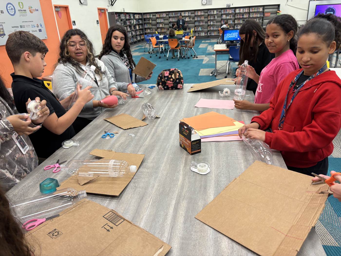 Students around a long table cutting cardboard fins and taping them to plastic bottles during a build session.